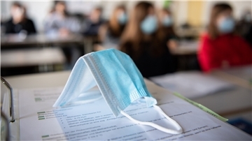 Eine Maske liegt im Unterricht auf Unterlagen, während im Hintergrund Schülerinnen und Schüler in einem Gymnasium mit Mund- und Nasenschutz zu sehen sind. Für Kinder ab 12 könnten Impfungen dieses Bild verhindern.