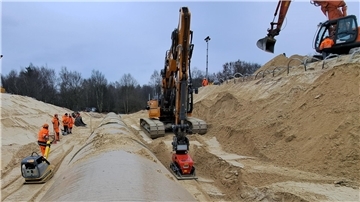 Eine Rüttelplatte wird von einem Bauarbeiter gezogen, die andere von einem Bagger: Schicht für Schicht wird der Sand um das neue Rohr verdichtet.
