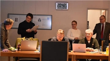 Personen sitzen am Tisch vor ihren Laptops, weitere Personen stehen im Raum.