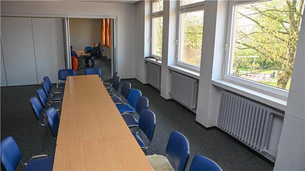 Wie wird das „Haus der Begegnung“ seinem Namen gerecht? Ein renovierter Raum mit aneinander gestellten Tischen und blauen Stühlen. Ein Baum taucht hinter einem Fenster auf.