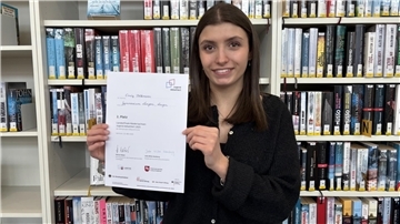 Emely Volkmann hält ihre Landessieger-Urkunde in die Kamera und lächelt. Im Hintergrund sind Bücher im Bücherregal zu sehen. 