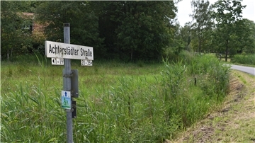 Straßenschild auf dem Achterstädter Straße zu lesen ist, viel grün und rechts ein bisschen Straße