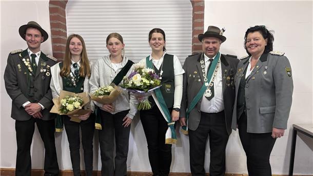 Sechs Mitglieder eines Schützenvereins stehen nebeneinander vor einer weißen Wand mit Ziegelbögen. Sie tragen traditionelle Schützenuniformen mit Orden und Schärpen. Drei Frauen in der Mitte halten Blumensträuße. 