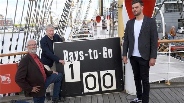 Drohnenshow und Schiffsparty: In 100 Tagen lockt die Sail Bremerhaven Drei Männer auf einem Schiff halten ein Schild in der Hand, auf dem „Days to go 100“ steht.
