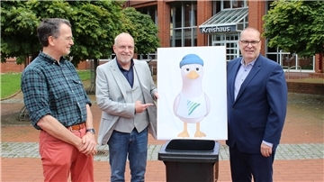 Vor dem Kreishaus stehen drei Männer neben einer Tafel mit der Zeichnung einer Möwe im blauen Mützchen. Die Figur ist Teil einer Abfallkampagne und lehnt an einer schwarzen Mülltonne.