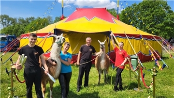 Eine Familie steht mit zwei Lamas auf einer Wiese vor einem gelb-roten Zirkuszelt. 