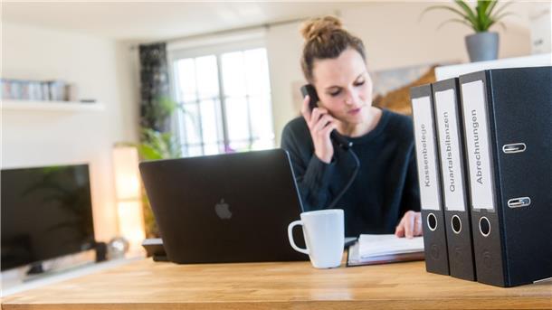 Eine junge Frau sitzt an einem Schreibtisch hinter einem Laptop und Aktenordnern, sie hält einen Telefonhörer ans Ohr und liest parallel in einem geöffneten Aktenordner