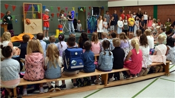 Viele Grundschüler sitzen im Halbrund auf Bänken in einer Turnhalle und wenden der Kamera den Rücken zu. Sie schauen auf eine Bühne, auf der ältere Kinder etwas vorspielen.