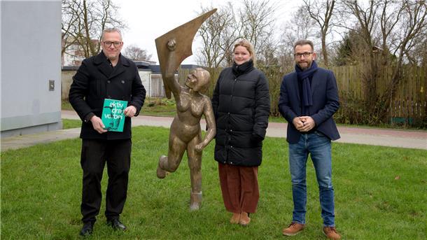 „Pauline“ lässt Drachen steigen: Gewoba schenkt Lehe ein neues Kunstwerk Drei Personen stehen auf einer Grünfläche vor einer lebensgroßen Bronzeskulptur, die eine Frau beim Drachensteigen darstellt.