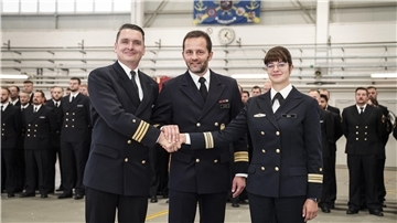 Bei einer feierlichen Kommandoübergabe stehen drei Marineoffiziere in Uniform in der Mitte eines Hangars. Zwei reichen sich die Hände, der Dritte legt seine Hand darüber. Im Hintergrund stehen uniformierte Soldaten in Reih und Glied.