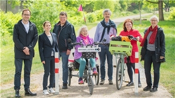Eine Gruppe von sieben Erwachsenen steht auf einem Rad- und Spazierweg im Grünen. Die Frau in der Mitte sitzt auf einem Dreiradtandem mit Schild "Abstand 1,50 Meter". Rechts neben ihr steht eine Frau mit rotem Mantel und Fahrrad mit grünem Transportkorb, in dem ein kleiner Hund sitzt.