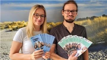 Eine junge Frau und ein Mann lächeln in die Kamera. In ihren Händen halten sie Flyer 