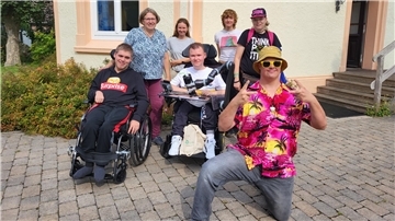 Sieben Menschen mit und ohne Beeinträchtigung stehen vor einem Haus und lächeln in die Kamera. 