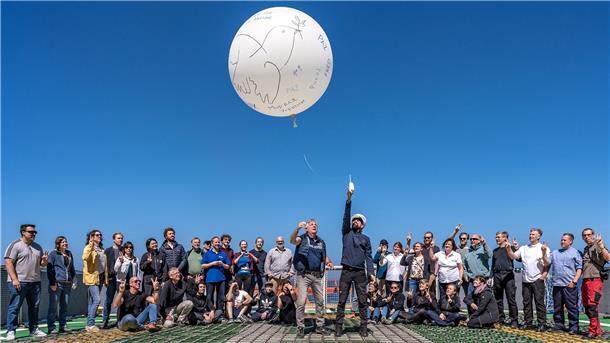 Mehrere Menschen stehen zusammen und deuten in Richtung Himmel. Dort fliegt ein großer weißer Luftballon.