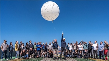 Mehrere Menschen stehen zusammen und deuten in Richtung Himmel. Dort fliegt ein großer weißer Luftballon.
