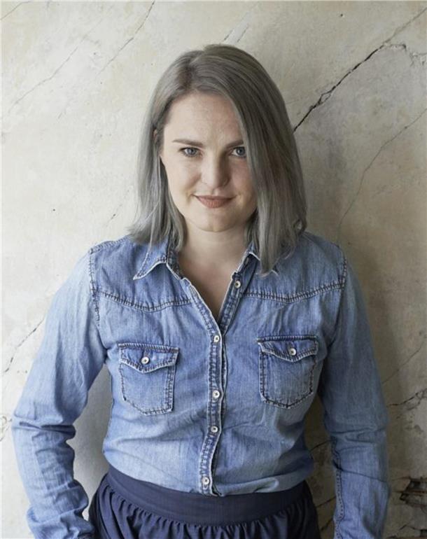 Masche für Masche: Friederike „Fredda“ Hansmann liest in Zeven Eine Frau mit halblangen Haaren in einem Jeanshemd steht vor einer Wand.