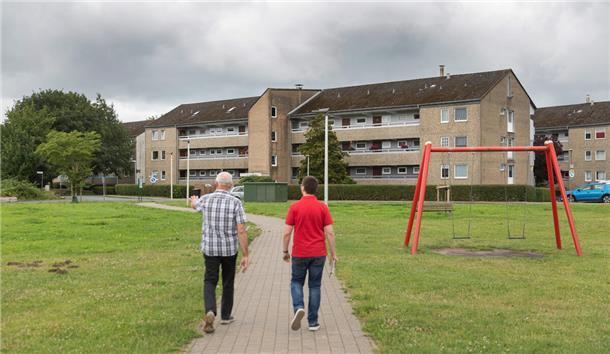Mehr Polizeipräsenz im „Brennpunkt Hohes Feld“? Was die Stoteler sagen Zwei Männer mit kariertem Hemd bzw. rotem Polo-Hemd sind von hinten zu sehen. Sie durchschreiten einen Park mit roter Schaukel. Vor ihnen wartet ein Häuserblock.
