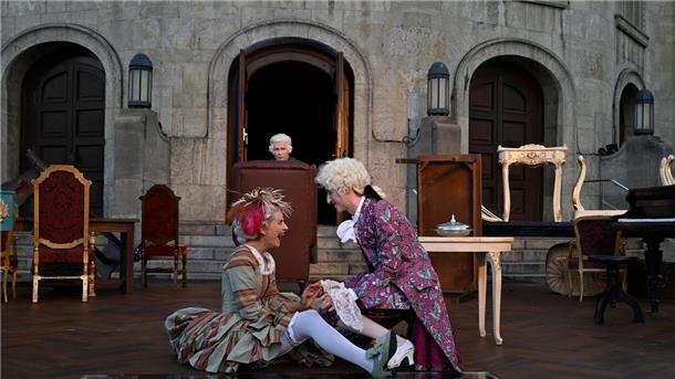 Auf einer Theaterbühne drei Personen in barocken Kostümen. Eine Frau und ein Mann im Vordergrund albern herum, im Hintergrund ein Mann im Ohrensessel.