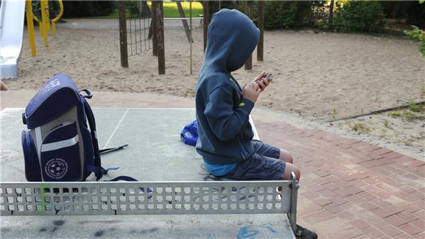 Ein Kind sitzt auf einer Tischtennisplatte und spielt mit dem Mobiltelefon. Daneben liegt der Schulranzen.