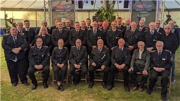 Gruppenfoto der Freiwilligen Feuerwehr Kirchwistedt anlässlich des 105-jährigen Jubiläums. Etwa 30 Feuerwehrmitglieder in Uniform sowie einige ältere Herren sitzen und stehen in mehreren Reihen in einem Festzelt. Im Hintergrund sind Bühnenelemente und Banner der Veranstaltungstechnik „Sound & Light Ringstedt“ zu sehen.