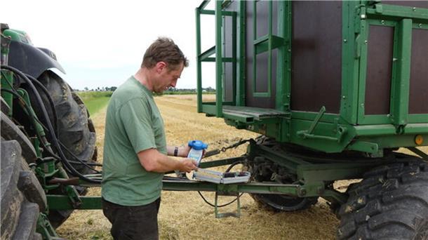 Ein Landwirt steht zwischen Traktor und Anhänger. Er hält ein Messgerät in Händen.
