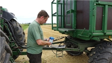 Ein Landwirt steht zwischen Traktor und Anhänger. Er hält ein Messgerät in Händen.