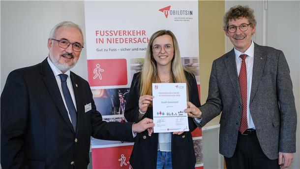 **ALT-Text:**  
Drei Personen bei der Urkundenübergabe zum Fußverkehrs-Check Niedersachsen; eine Frau in der Mitte hält die Urkunde der Stadt Geestland, flankiert von zwei Männern im Anzug, im Hintergrund ein Roll-up mit der Aufschrift „Fußverkehr in Niedersachsen“.