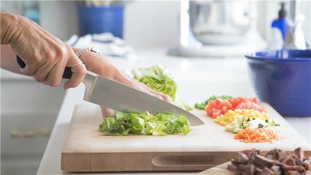 Auf einem Holzbrett wird Salat mit einem großen Messer geschnitten. Im Hintergrund stehen Küchenutensilien.