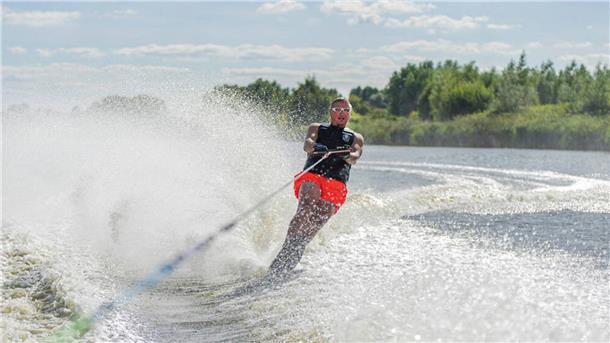 Ein Mann läuft Wasserski. Dabei trägt er eine orangefarbene Badehose und eine schwarze Schutzweste, sowie eine Schwimmbrille.