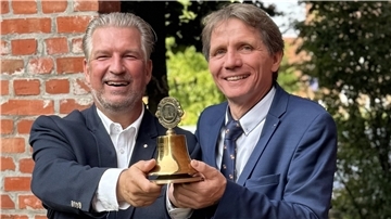 Jörg-Stephan Garms und Uwe Krüger stehen nebeneinander, lachen und halten gemeinsam eine goldene Lions-Club-Glocke, im Hintergrund eine Backsteinmauer und Bäume.