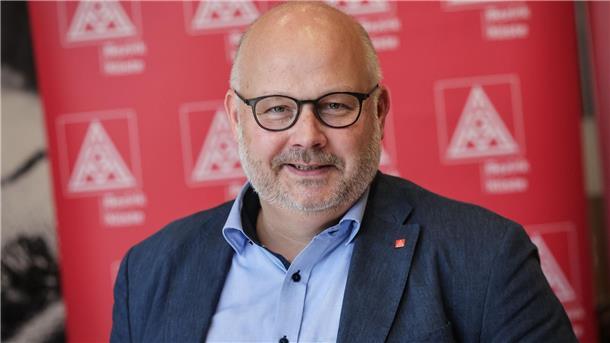 Porträt eines Mannes mittleren Alters mit Brille, hellem Haarkranz und kurzem Bart. Er trägt ein blaues Hemd und ein dunkles Sakko. Im Hintergrund ist ein roter Banner mit dem Logo der IG Metall zu sehen.