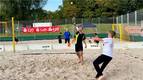 Zwei Männer spielen auf einem Beachfeld ein Spiel mit Schlägern und Ball. 