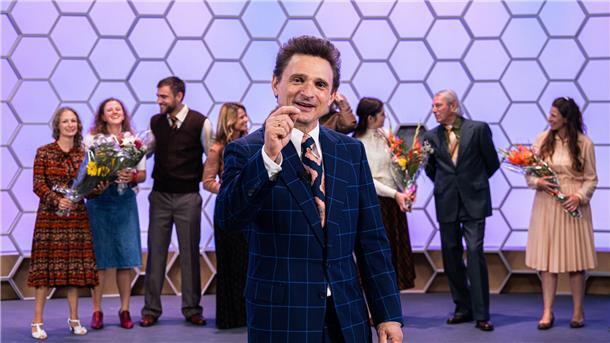 Hans Rosenthal (Florian Lukas) gestikuliert lächelnd im bunten "Dalli-Dalli"-Fernsehstudio. Hinter ihm stehen einige gut gelaunte Kandidatinnen und Kandidaten (Komparsen). Das biografische Drama „Rosenthal“ wird am Montag, 7. April (20.15 Uhr), im ZDF gezeigt.