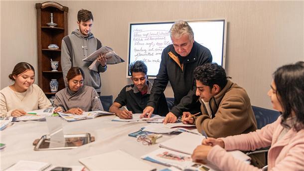 Ein Lehrer steht an einem Tisch, um den herum sechs Schüler sind. Der Lehrer zeigt ihnen etwas in einem Arbeitsbuch.