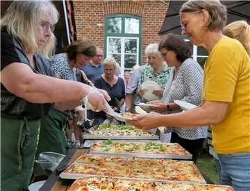Eine Frau gibt Pizza von großen Backblechen aus. Andere Gäste warten mit Tellern in der Hand. 