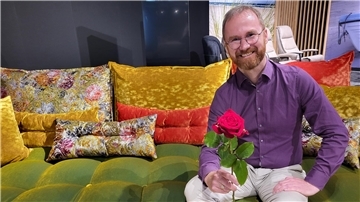 Ein Mann sitzt lächelnd auf einem bunten Sofa und hält eine Rose in der Hand. 
