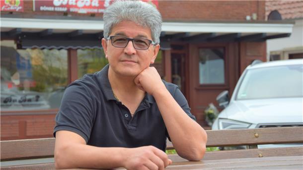 Es ist ein Herr in einem kurzärmeligen schwarzen Polo-Hemd zu sehen. Ibrahim Dogan stützt seinen Kopf auf seiner linken Faust ab. Im Hintergrund ist sein kürzlich eröffneter Imbiss Baba's zu sehen.  