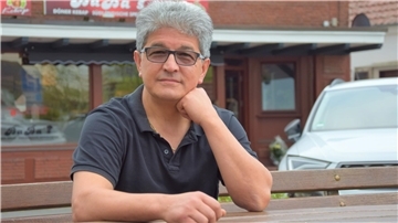 Es ist ein Herr in einem kurzärmeligen schwarzen Polo-Hemd zu sehen. Ibrahim Dogan stützt seinen Kopf auf seiner linken Faust ab. Im Hintergrund ist sein kürzlich eröffneter Imbiss Baba's zu sehen.  
