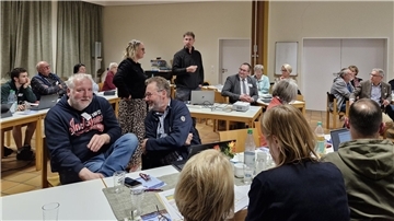 Menschen sitzen in Gruppen an Tischen in einem Konferenzraum und diskutieren lebhaft während einer Sitzung. Einige Teilnehmende arbeiten an Laptops, andere stehen oder unterhalten sich. Auf den Tischen liegen Getränke, Unterlagen und Namensschilder.