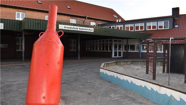Ansicht des Schulhofs der Grundschule Rodenkirchen mit einem roten maritimen Dekorationselement im Vordergrund. Im Hintergrund ist das Backsteingebäude mit rotem Dach und grüner Überdachung zu sehen. Rechts befindet sich ein Spielbereich mit Klettergerüst und Sandkasten.