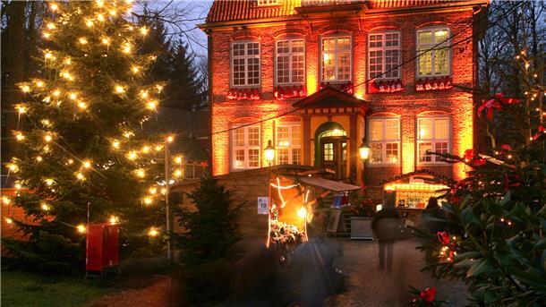 Beleuchtetes Schloss mit geschmückten Tannen davor, sowie zwei Holzbuden.
