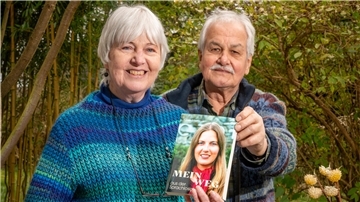 Ein älteres Ehepaar lächelt in die Kamera und hält ein Buch in der Hand mit dem Titel „Mein Weg aus der Sprachlosigkeit“.