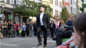 Ein Mann geht auf einer Stra0e - scheinbar, beobachtet von vielen Menschen. Ist das noch die Straße oder doch ein Turnierplatz? Paul Hess verwandelt sich in ein Pferd, nicht in irgendeines, sondern in den Wunderhengst Totila, der mehrere Goldmedaillen gewann.