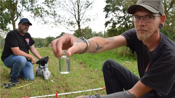 Ein Mann hält ein Gläschen mit Wasser in die Kamera, daneben hockt ein zweiter Mann mit Hund.