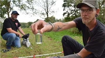 Ein Mann hält ein Gläschen mit Wasser in die Kamera, daneben hockt ein zweiter Mann mit Hund.