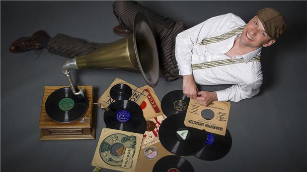 Ein Mann liegt lachend neben einem Grammophon am Boden. Neben ihm liegen Schallplatten. 