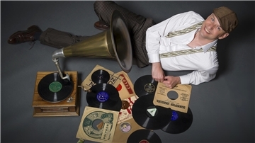 Ein Mann liegt lachend neben einem Grammophon am Boden. Neben ihm liegen Schallplatten. 