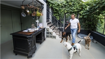Jochen Bendel in einem Wintergarten-ähnlichen Raum mit Pflanzen und drei Hunden. Der Raum wirkt einladend mit einer rustikalen Einrichtung und dekorativen Elementen.