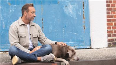 Jochen Bendel sitzt im Schneidersitz vor einer blauen Tür und legt seine Hand auf einen Labrador, der neben ihm liegt. In der Arbeit mit Hunden hat Moderator Bendel seinen Traumberuf und seine Berufung gefunden.