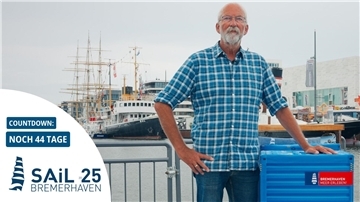Ein Mann mit kariertem Hemd steht am Hafen von Bremerhaven vor Segelschiffen. Im Bild ist ein Countdown zur Sail 2025 mit „Noch 44 Tage“ eingeblendet.